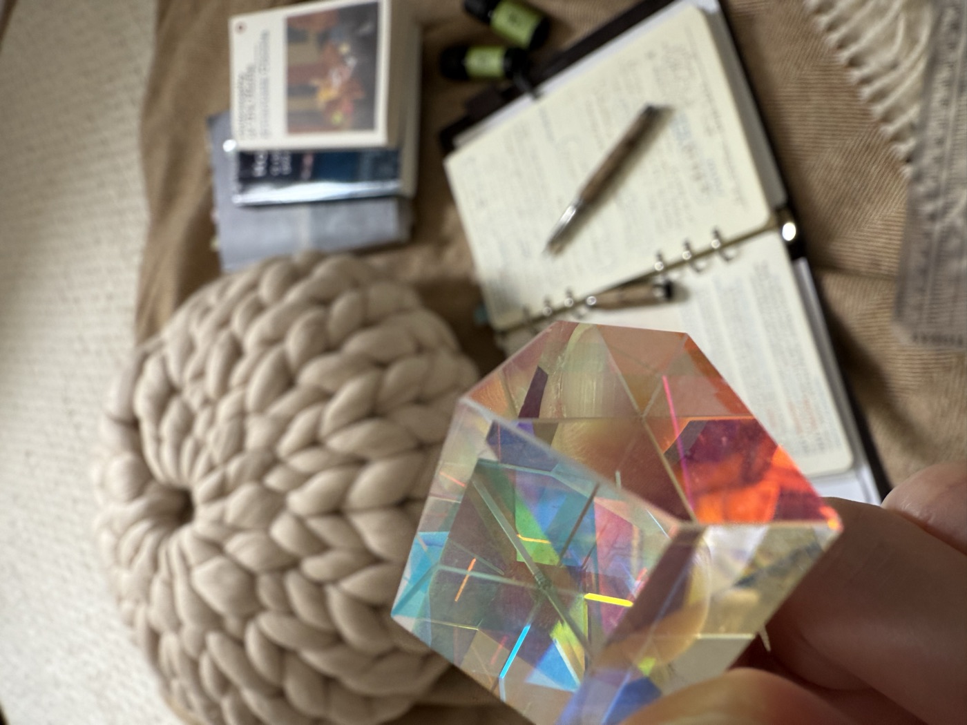A small glass house held up to the light, over an open notebook, a pen, a stack of books and two bottles of oil on a knitted throw.
