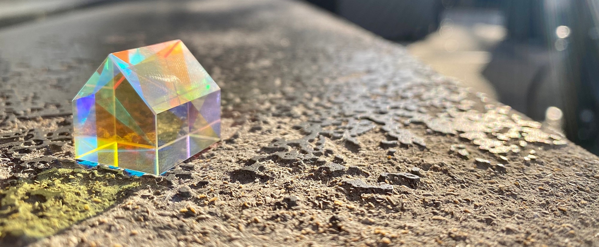 A small glass house on a sunlit stone ledge, its sides holding bands of coloured light.